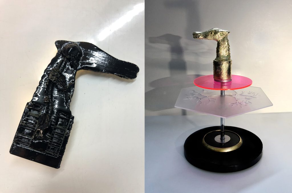 Photography: Different layered objects are held from a metallic bar. The bar  is standing on a round black pedestal and has at the top the golden head of a canine-like deity. Under this 3D printed object there is a circular pink acrylic plastic disk, followed under, and in the middle of the metallic bar, a decorated translucent acrylic plastic with an hexagonal shape. On a second image, a black canine-like deity printed on 3D.