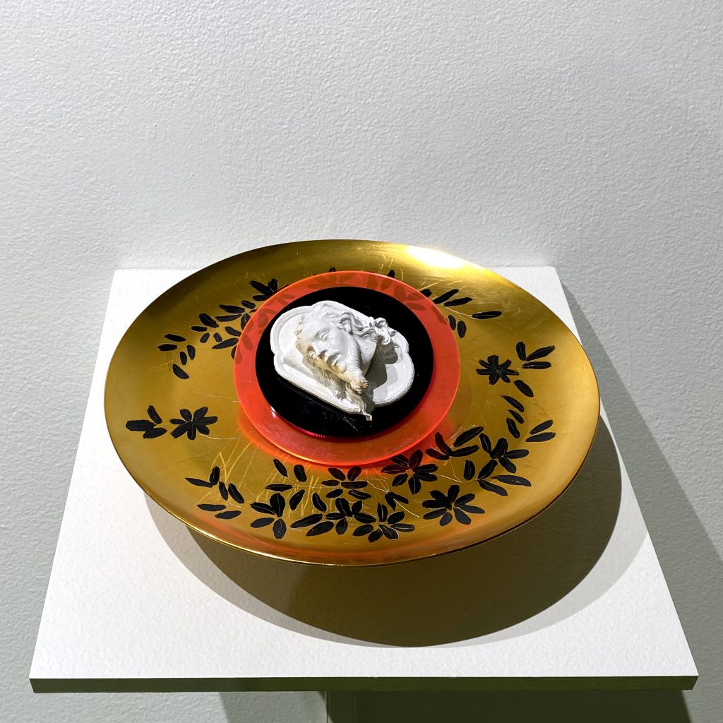 Photography: A golden tray decorated with black floral motifs on a white shelf. In the center of the plate, two overlapping circles. The first translucent phosphorescent pink one made of acrylic plastic and a second smaller black one on which is a small white sculpture of a male face with beard and white hair.