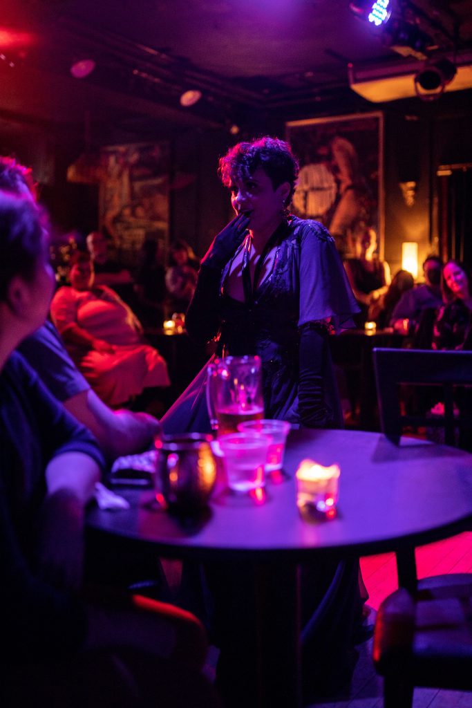 Image: Tila Von Twirl performing at a club, walking among the audience, placing a hand to her mouth. The lighting of the nightclub is purple and the audience watches Tila from their tables enraptured. In the foreground is a table with drinks and a candle on it. Photo by Erica McKeehen.
