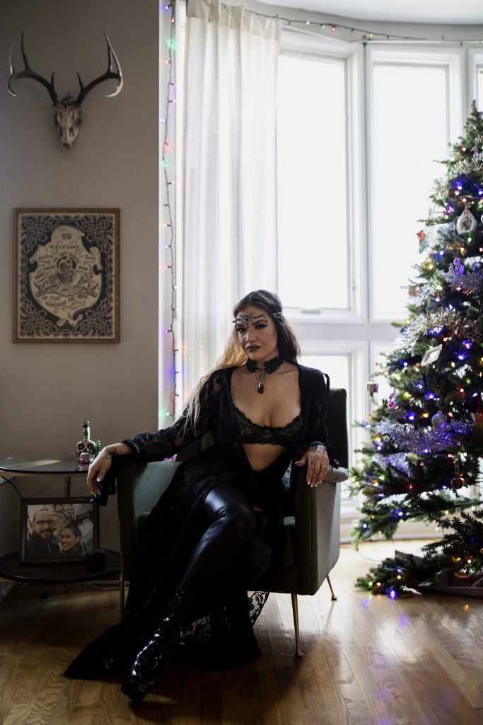 Image: Miss Nyxon sits on a black chair in her living room looking towards the camera. There is a decorated Christmas tree to her left and a family photo to her left on an end table. She is dressed in costume, wearing all black. Photo by Erica Mckeehan.