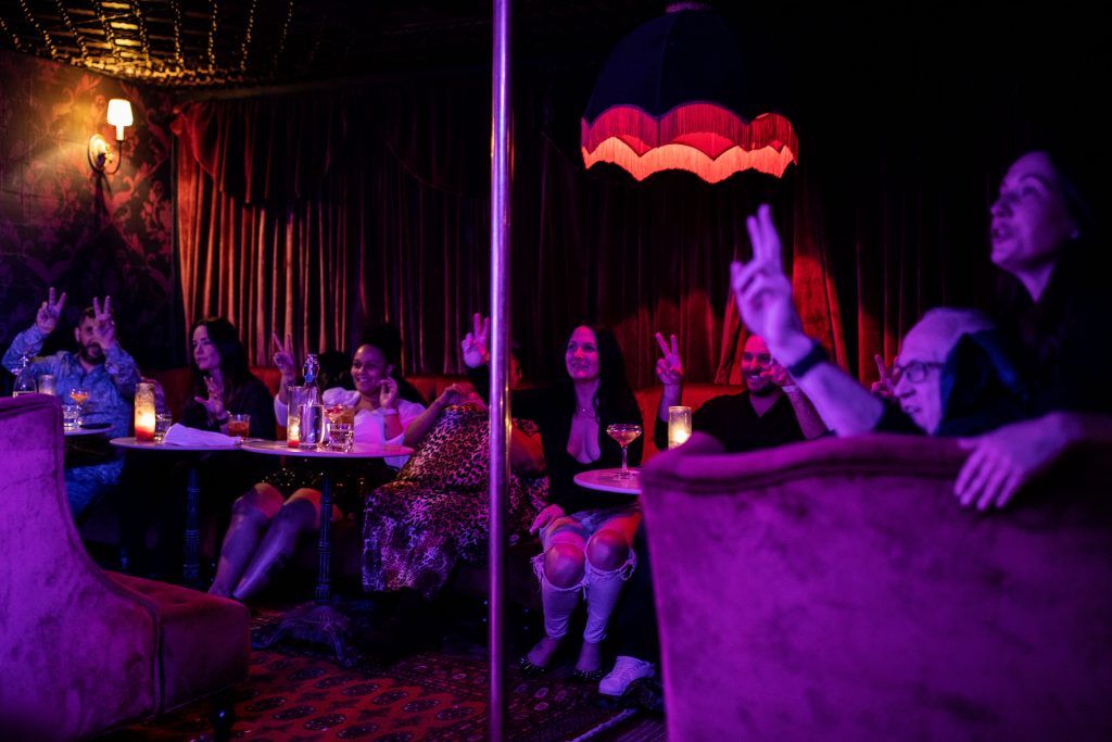 Image: Audience members get into the spirit before the show begins at Bordel, 2022. About ten people bathed in purple and pink light and seated in plush drink booths, hold up peace signs. There is a metal pole dividing the image. Photo by Erica McKeehen.