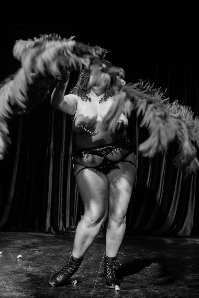 Image: Camille Leon disappears in a flurry of her feather fans during The Shakin’ It Show at The Newport Theater, 2022. The feathers extend toward the image, obscuring her face. She wears pasties with tassels and black boots. Photo by Erica McKeehen.