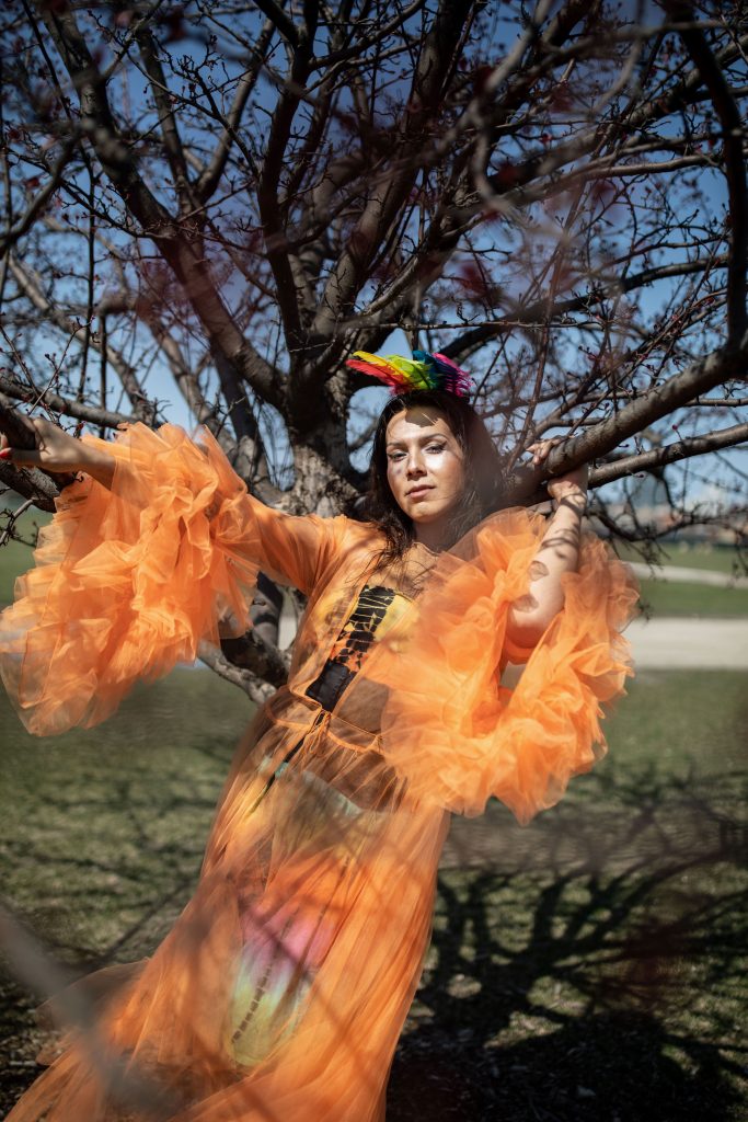 Image: Portrait of Mz Mr, “Princess of Pilsen,” taken in 2023 at a park in front of a tree in Chicago. They are leaning on the tree and wearing a rainbow headpiece, a light orange sheer dress with oversized ruffled sleeves with a colored dress underneath. The dress is flowing in the wind. Photo by Erica McKeehen.