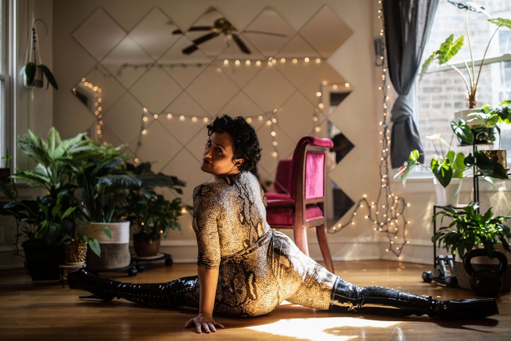 Image: Tila Von Twirl lying in the splits facing their body away from the camera and turning their head towards it. They wear black boots and a form-fitting snakeskin print "catsuit. Surrounding them are plants. Directly behind them is a velveteen, red chair and a grid of mirrors that reflects the stringlights on an otherwise unseen wall. Photo by Erica McKeehen.