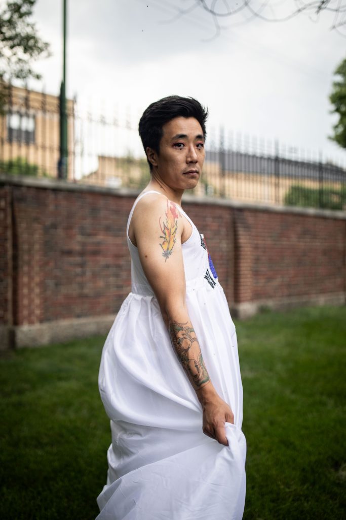 Image: Joona Bae, 2023. Joona Bae is pictured faced right with their head looking towards the camera. Joona is standing in a grassy field wearing a long, white, tank-top sleeved, flowy dress with colorful embroidery on the chest section. Joona has colorful tattoos on their right arm. Photo by Erica McKeehen.