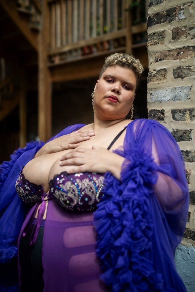 Image: Highness Noire taken in 2023. Highness Noire is wearing a purple sheer tulle robe, sequined and sheer camisole with a ribbon in the center of their chest between their breasts. They stand outside, near a brick wall and a wooden patio. Photo by Erica McKeehen.