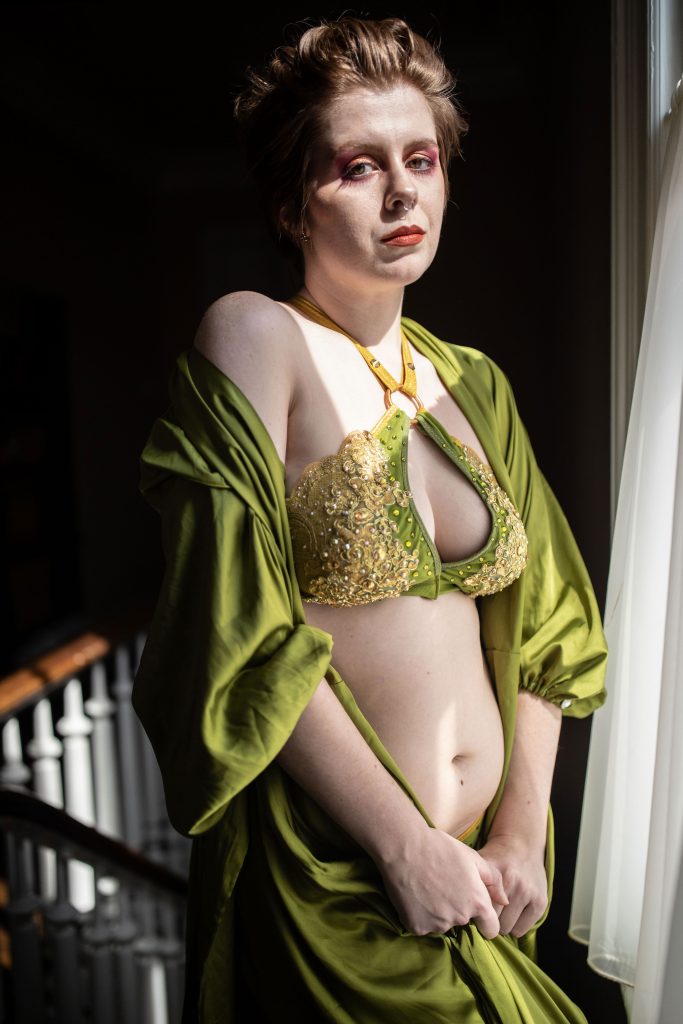 Image: Kevlar B Lightning. Lightning is wearing a green sequined and lace bra, and a matching green cloak. They stand in a stairwell, looking at the camera with pink eyeshadow and orange lipstick. They clutch their cloak just beneath their belly. Photo by Erica Mckeehen.
