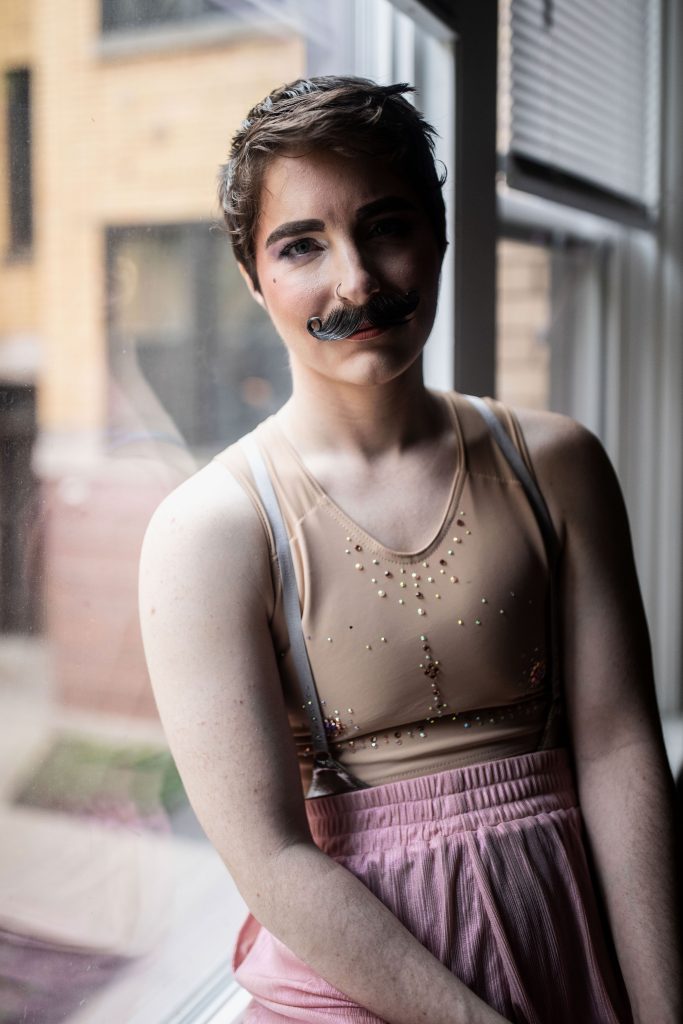 Image: Portrait of Matt Adore in 2023. Adore has brown hair and a handlebar mustache. They are wearing silver suspenders, pink bottoms and a flesh-colored/nude tanktop with colored sequins around their chest. They pose in front of a window with an apartment building in the background. Photo by Erica Mckeehen.