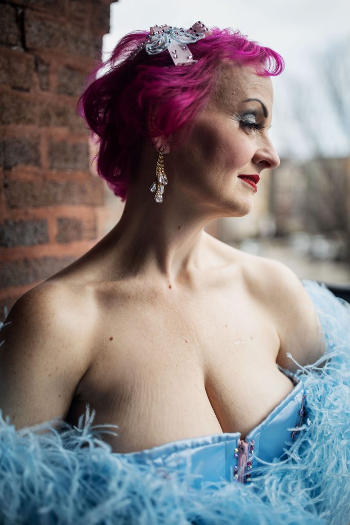 Image: A right-side profile image of Lady Ginger, 2023. Lady Ginger is photographed in her burlesque costume and makeup, which includes a light blue corset and matching feathered boa. She has hot pink hair and is wearing chandelier makeup. Photo by Erica McKeehan.
