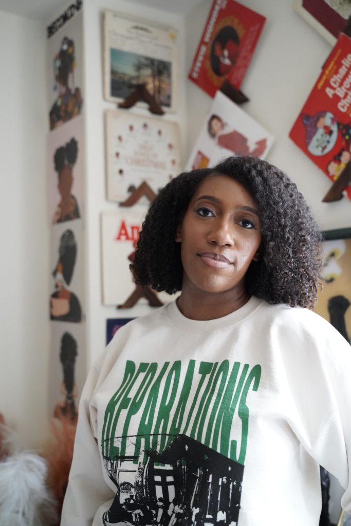  Image: A portrait of Redmond in a white sweater with the top half of the word “REPARATIONS” printed in green text on the shirt. Behind her are various prints and art pieces hung on white walls. Photo of Jalen Hamilton.