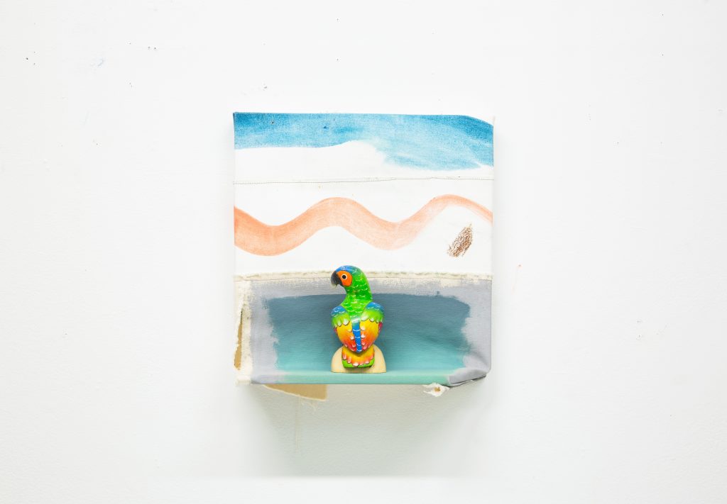 Photography: square shaped small Unframed painting with four colors painted across (blue, white, orange) giving a sense of horizon and the beach. The lower part of the painting becomes a small shelf. Standing on the shelf, a small, green, red, and yellow parrot. The second image is a different angle of the work.