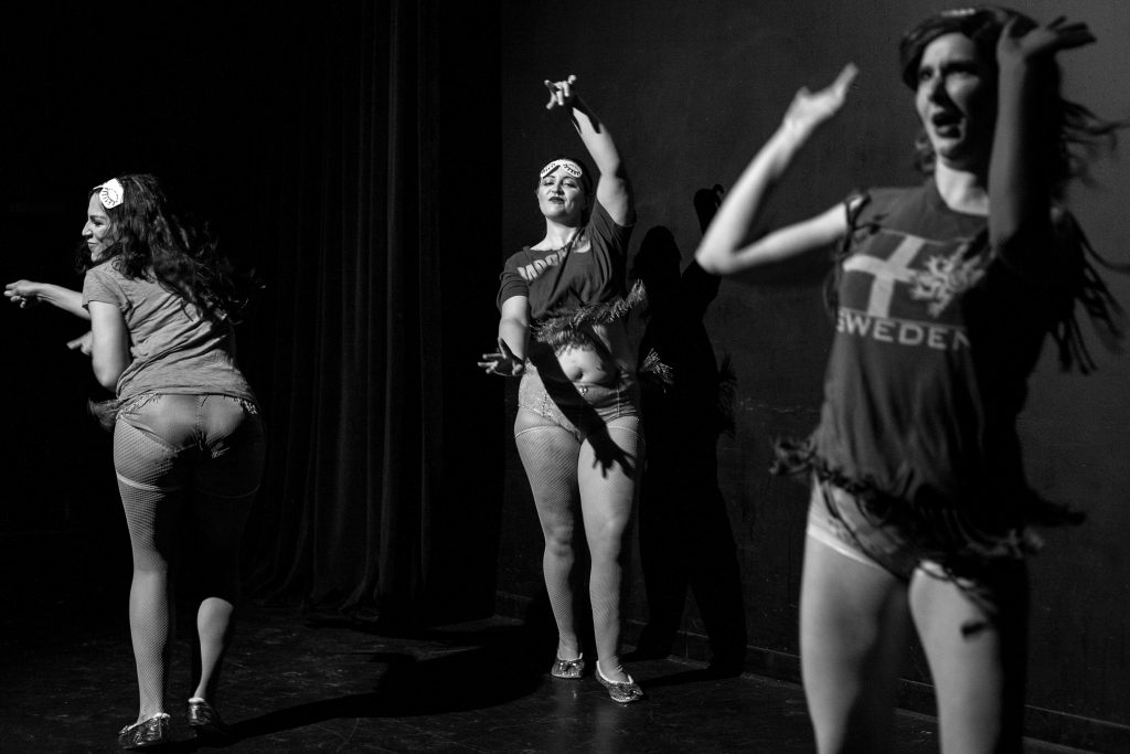 Image: A black and white image of Valencia Vice, Kitty Tornado, and Secret Mermaid performing a group act in our formal ensemble at Stage 773, 2018. All three performers are pantsless, wearing sleep masks on their foreheads, and in various state of active arm gesture. Photo by Erica McKeehen.