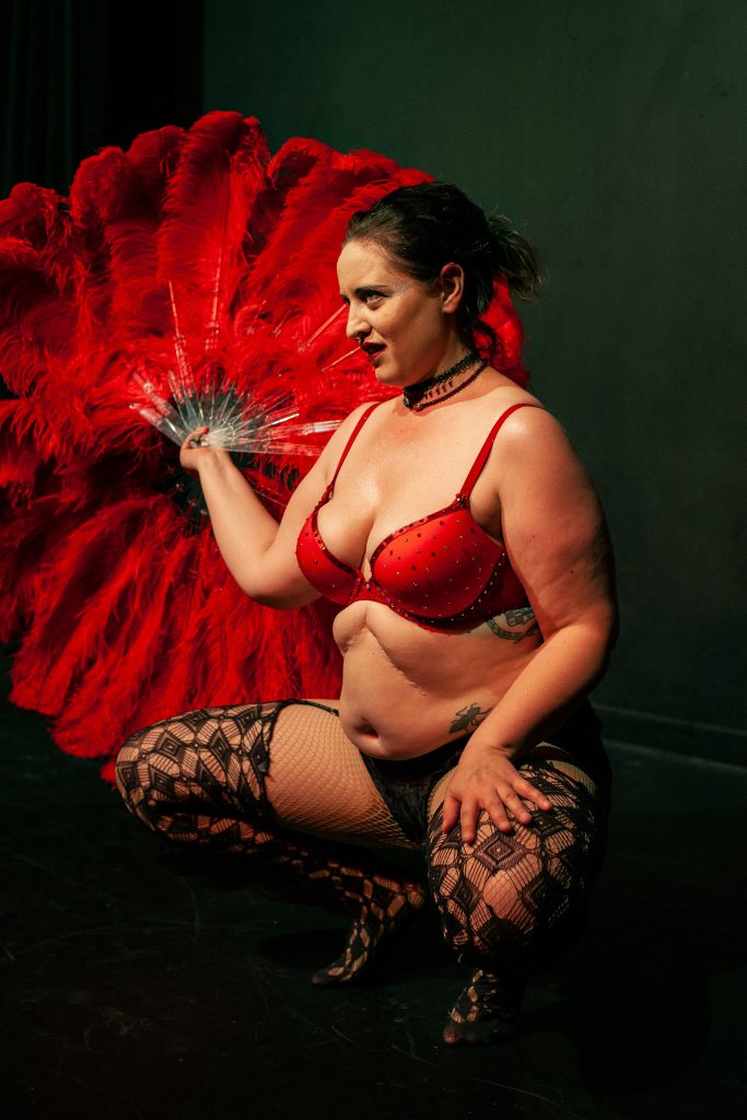 Image: Kitty Tornado's “Only Sour Cherry” fan act, 2018. Kitty Tornado is pictured facing towards the left of the camera wearing a red bra, black patterned tights and a choker necklace. They are holding a red feather fan in their right hand. Photo by Erica McKeehen.