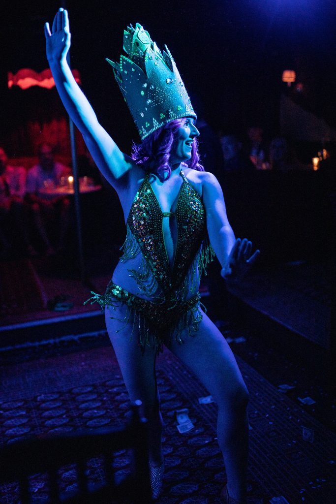 Image: Secret Mermaid performs her “Ass-paragus” act during Vaudeville at Bordel, 2022. Secret Mermaid has pink hair and is wearing a tall bejeweled crown and bathing-suit like bejeweled lingerie. The light on them is a cerulean blue. Photo by Erica McKeehen.