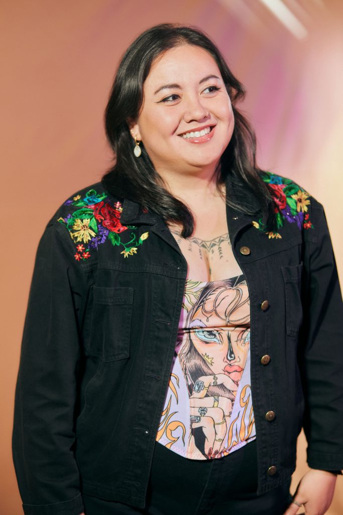Image: Poet Chris Aldana. She's standing in front of an orange back drop, she gazes to the right with a smile on her face She wears a black jacket with colorful flowers on the shoulders. Photo by Sarah Joyce
