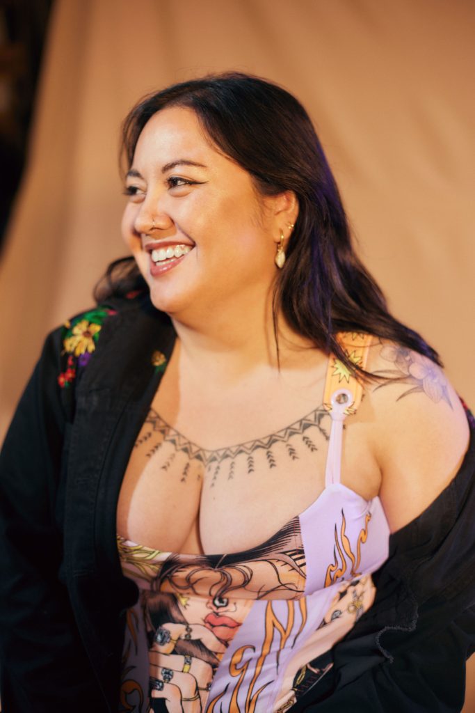 Image: Poet Chris Aldana. She's seated, looking toward the left, and she's smiling broadly. Her hair is pushed behind her ear, showing a series of dangling earrings. Her jacket is falling off her shoulder. Photo by Sarah Joyce