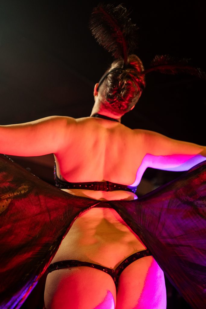 Image: Helena Handbasket reveals their rendition of the Mothman during Lust for Life at Dorothy, 2023. Helena Handbasket, facing away from the camera is wearing a moth-like lingerie set equipped with black wings and a moth-antennae headpiece. Photo by Erica Mckeehen.