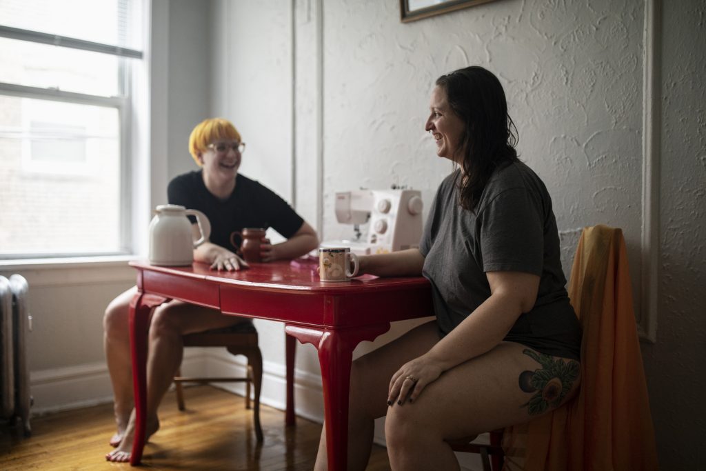  Image: Kitty Tornado and I have been best friends and creative collaborators since meeting in 2014, 2022. Kitty Tornado and the author, Erica McKeehen, pictured holding coffee mugs and laughing mid-conversation at a red dining table in Kitty Tornado's apartment. Both Tornado and Mckeehen are wearing a black t-shirt and no bottoms. A sewing machine is at the center of the table between them. Photo by Erica McKeehen.