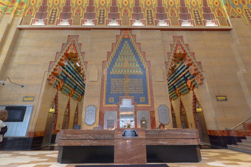 Image: Lobby of the Guardian Building in Detroit, MI. Art Deco tiles in greens, blues, ochres, and dark corals, rise upwards on high walls. A large stone lobby security desk site in the middle of the photograph, behind the desk are six elevators; three in each alcove separated by the large stone support that sites behind the desk. A staircase ascends out of frame. A person sits behind the desk as another person stands left of center and looks up. Photo by Mikey Mosher.