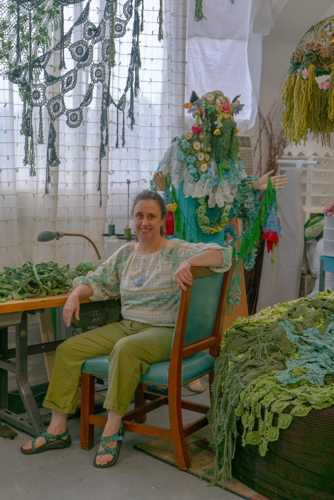 Image: Melissa Webb in her studio. The artist wears sandals, lime green pants, and a patterned blouse in a large bottle green and wooden hard back chair. She sits by a desk covered in crocheted green fabric. A mannequin covered in colorful crocheted fabric, stands behind the artist. A gauzy curtain obscures the window behind the desk. Against the studio's white walls ringlets of crocheted fabric rain down to the floor and a large pile of fabric in various shades of greens sits to the right of the artist. Photo by Mikey Mosher