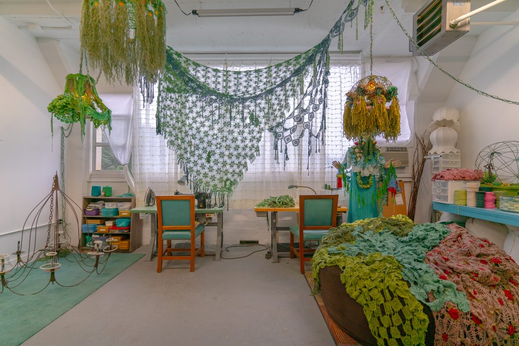 Image: Melissa Webb's artist's studio. A wire candelabra sits on top of a green rug. Above the light fixture hangs a curving, naturalistic lamp covered in crocheted green fabric. Windows covered by a gauzy curtain line the studio's back wall. Two desks are pushed against the back wall, each with a similar green and wooden chair. A bookshelf of Tupperware cases stands to the left of the desk, and to the right is a mannequin draped with fabrics of various hues and weaves. In front of the mannequin is a large cushion covered with woven pink, blue, and green fabrics. Above the draped cushion, various threads are stacked on a blue shelf. Photo by Mikey Mosher.