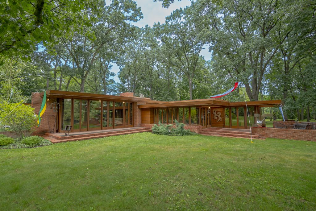 Image: Exterior of Frank Lloyd Wright's Smith House. A brick house built in the style of mid-century Modernism diagonally extends across a green lawn. The house's mirrored windows reflect the surrounding trees. A green and yellow flag hangs to the left of the house and a red and white flag flies on a flagpole to the right of a stone porch. Photo by Mikey Mosher