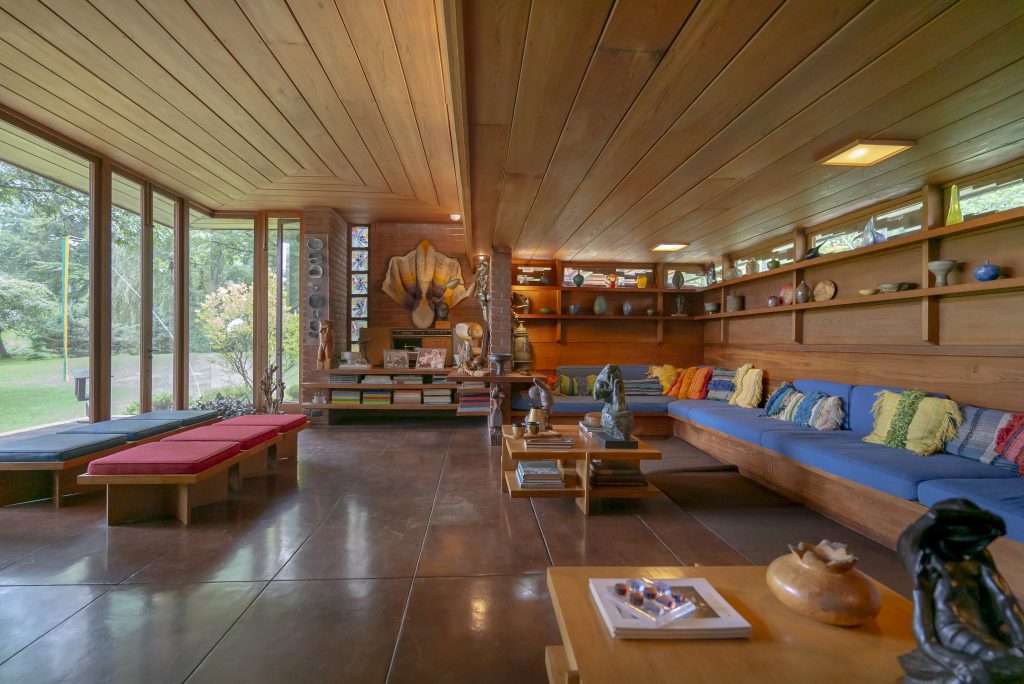 Image: Living room of Frank Lloyd Wright’s Smith House. Wooden beams run in parallel lines across the ceiling as dark tile matches their pattern on the floor. Against large floor to ceiling windows muted blue and red seats are arranged. Books and art populate the space's many shelves and a wrap-around blue couch faces the windows. Photo by Mikey Mosher