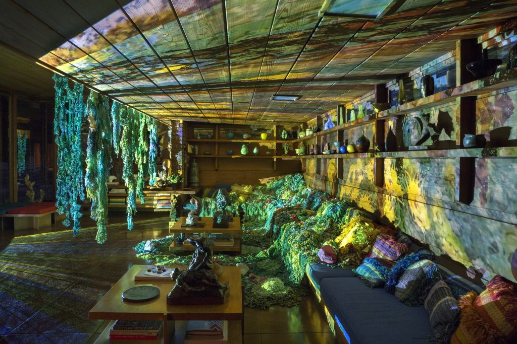 Image: Installation view of local authorities in the spirit world shape shift through time (we call it evolution) at night. Psychedelic light and distorted images of flowers and greenery illuminate the Smith House sitting area. The greens of Webb's hanging installation shift their tones against the light, as the work installed on the couch turns a watery yellow. The projection's source radiates outward, left of frame. Please reference the previous photo caption for all artist collaborators. Photo by P.D. Rearick. 