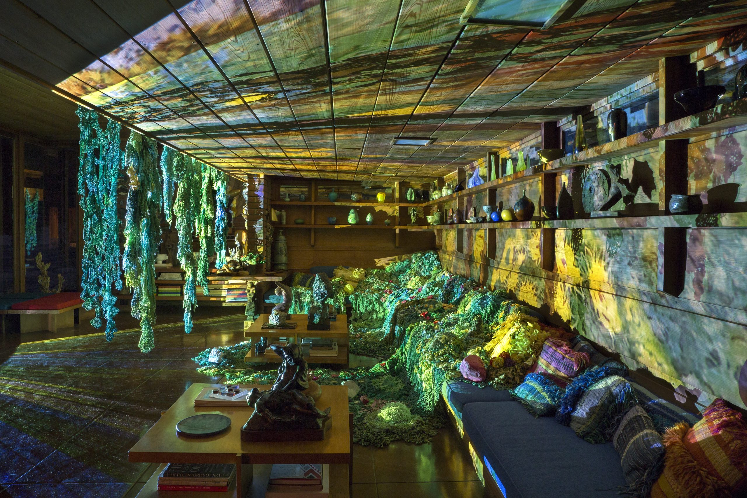 Image: Nighttime installation view of local authorities in the spirit world shape shift through time (we call it evolution) by Melissa Webb. Interior shot of Frank Loyd Wright designed home shows a sitting area bathed in psychadelic light. The many colored lights come from an artist's projection out of frame and cast green and yellow shadows upon the wooden walls. Textile pieces, made from crocheted fabric hang from the ceiling and the couch. Photo by P.D. Rearick.