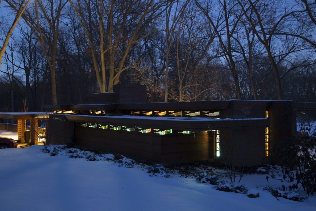 Image: Exterior view of local authorities in the spirit world shape shift through time (we call it evolution) at night. At night outside the Smith House, snow sits on the ground and the yellow light from the installation projection can be seen in pockets through the reflecting windows. Images, shapes, and colors can be made out near the tops of windows and trees near the back of the house are lit up with the projection's light. Please reference earlier image caption for all artist collaborators. Photo by P.D. Rearick and Melissa Webb.