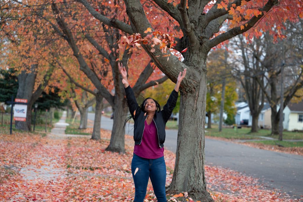 Image: On a suburban street lined with trees, Alyssa stands center with her arms up in the air and smiling, looking up at the arc of red and orange leaves she's just thrown in in the air. Behind her the lawns and sidewalk are littered with more fall leaves. To the right is a car-bare asphalt street. On the other side of the street, there are leafless trees and one story houses. Photo by Tonal Simmons.