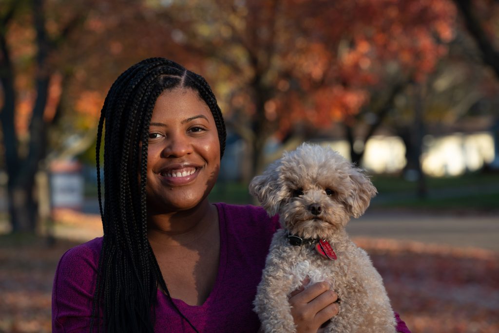 Image: Alyssa Laney from the chest up. Her braids fall over the right side of her face and down her shoulder. She is smiling broadly. In her left arm is a small and fuzzy light brown dog. Photo by Tonal Simmons.