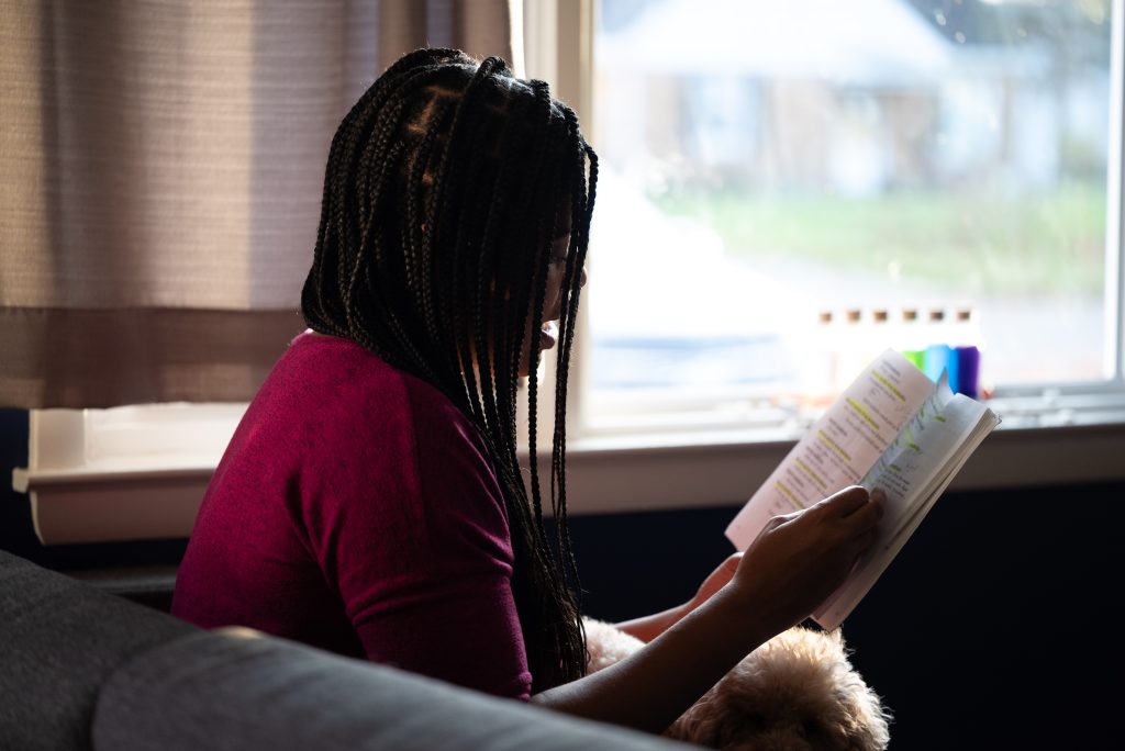 Image: Alyssa Laney seated in profile on a couch in front of a bright window. Her braids obscure her face slightly while she reads from an open book with lines of text highlighted. On the window sill are six bottles filled with vibrantly colored liquids. Her dog sits on her lap. Photo by Tonal Simmons.