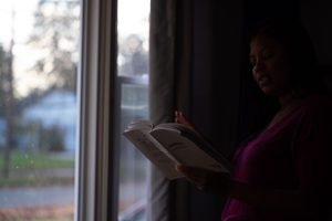Image: A photo of Alyssa Laney reading a white book. On the left side of the image are windows that go from the bottom of the image to the top. Outside, it looks to be dusk in a suburb. On the right side of the image, Alyssa—lit only by the window—holds a white book in front of her, raising her right hand slightly. She wears a purple shirt. Photo by Tonal Simmons.
