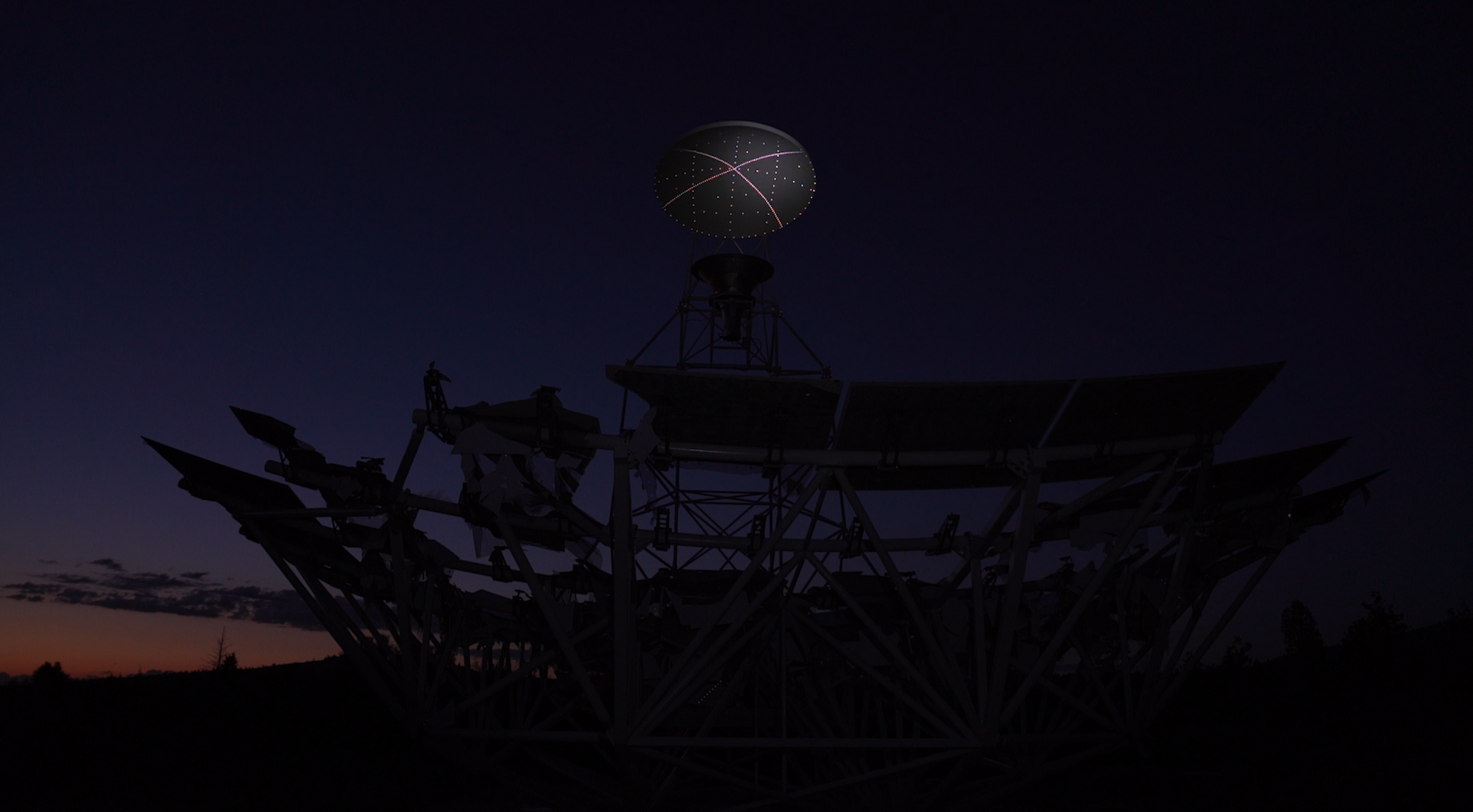 Image: Brittany Nelson, still frame from "I can't make you love me," single channel video, 2024. The Allen telescope, a many-faceted structure, is silhouetted in the foreground, much of its detail is hard to see. Behind it, is a barely lit horizon. Image courtesy of Patron Gallery.