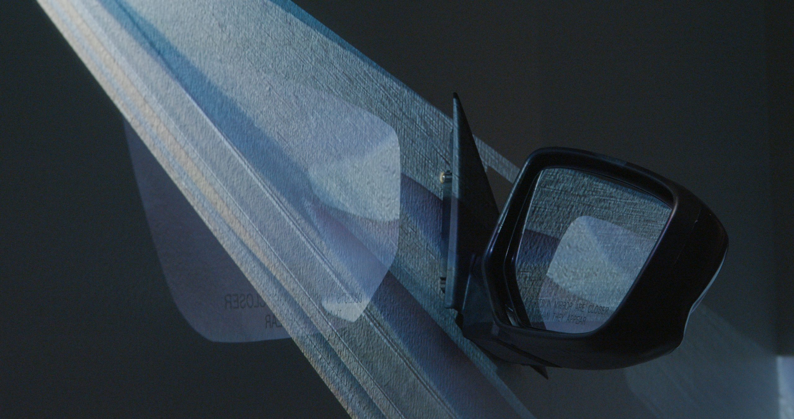Image: Ruby Que, Objects in Mirror are Closer than They Appear, 2024. A car's passenger side mirror is mounted to the wall. A projected video of bluish light bisects the image on a diagonal, starting in the upper left corner and ending in the bottom right. A reflection of the image is faintly visible on the wall. Photo by Ruby Que.