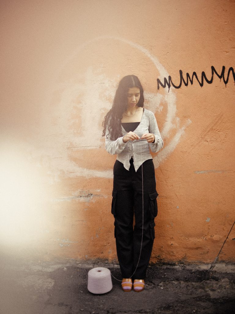 A photograph of Carolina Vélez Muñiz with a roll of yarn. She is wearing black pants, a cream colored shirt, and has her long curly brown hair wore down. Standing in front of an orange wall that has black graffiti on the right side of her face. 