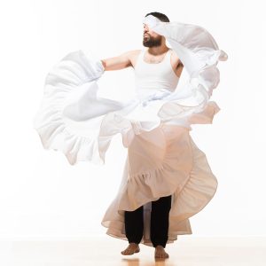 Image: A promotional photo from "World After This One," an experimental dance performance by Benji Hart. Hart is captured in movement, wearing a white tank top and skirt. The skirt is flung upward, covering half of their face. Photo by William Frederking.