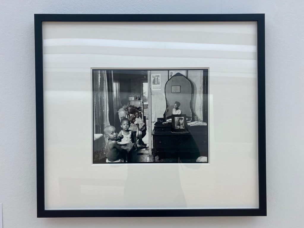 Image: A framed photo. Gordon Parks, Mrs. Ella Watson, a government charwoman and her grandchildren, August 1942. The inside of a home. On the left side of the image are, from foreground to background, a shirtless baby looking at the camera, a toddler facing the camera, an older woman looking at an object in her hands, then another child. There is a door leading directly outside. On the right side of the image, separated by a doorframe from the other figures, is a vanity bureau with a mirror reflecting a woman and a framed certificate above her head. Photo by Jasmine Barnes.