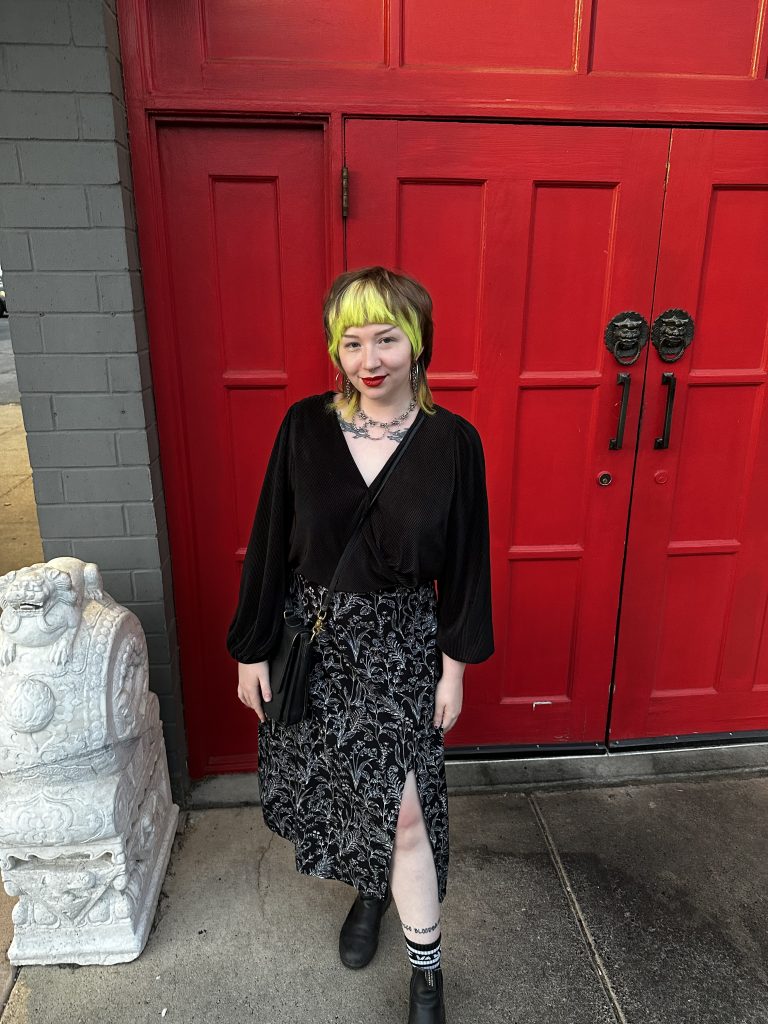 A photo of Summer who is standing in front of a red door and is wearing a black long sleave shirt and a black and white skirt. She has shoulder length green and brown hair.