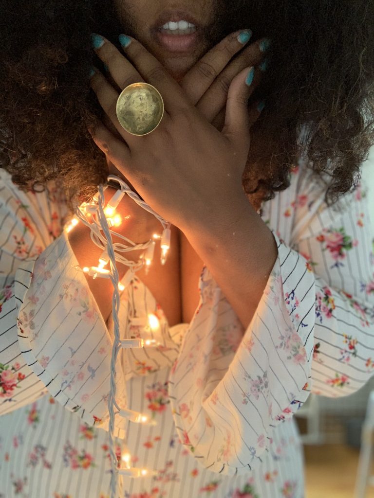 Image: Tonal, wearing a a striped white dress with a floral pattern, holds their overlapped hands over their throat. Their fingers frame their slightly open mouth. Draped down from their right hand are yellow LED Christmas lights. The light represents the loss of voice. Photo by Tonal Simmons.