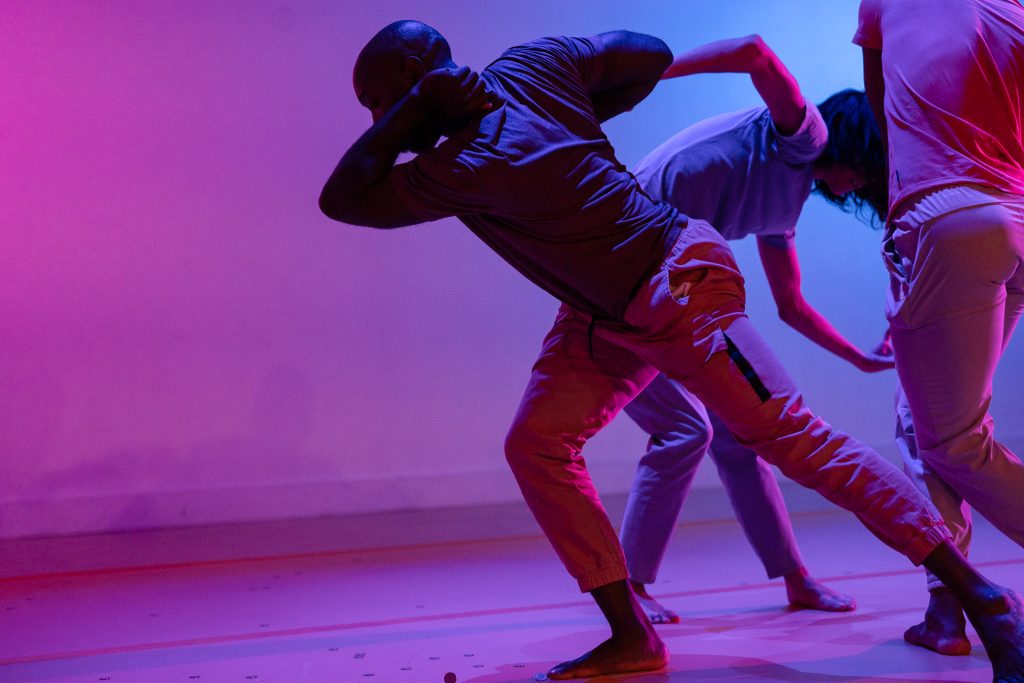 Image: A dancer lunges towards the left side of the image with his elbow jutting beyond his face. Behind him are two figures gesturing around each other. A bare wall to the left. Lights are pink, purple and blue. Photo by Michelle Reid.