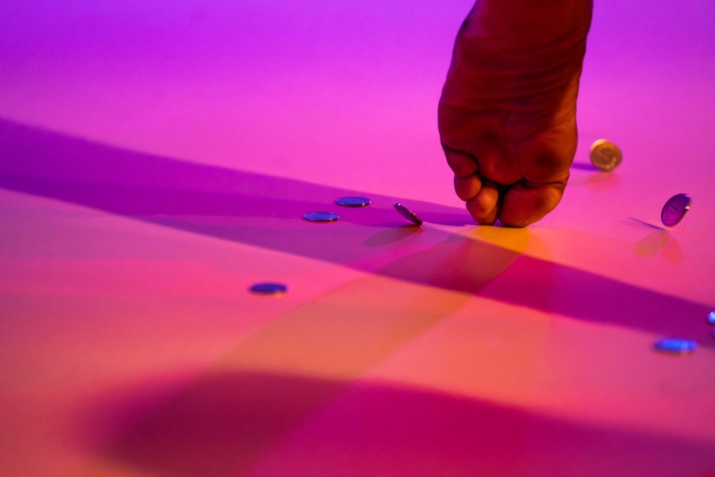 Image: On a purple, pink, and orange lit floor, the bottom of a foot faces the camera, its toes pressing into the floor. Surrounding it are several coins, some resting on the floor, others mid-bounce. Shadows cast from the foot spread in many directions. Photo by Michelle Reid.