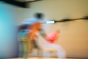 Image: A blurred still from "Righteous Beauty." Three dancers are seen in motion, one in the mid-ground, two in the background in a tan room. Photo by Michelle Reid.