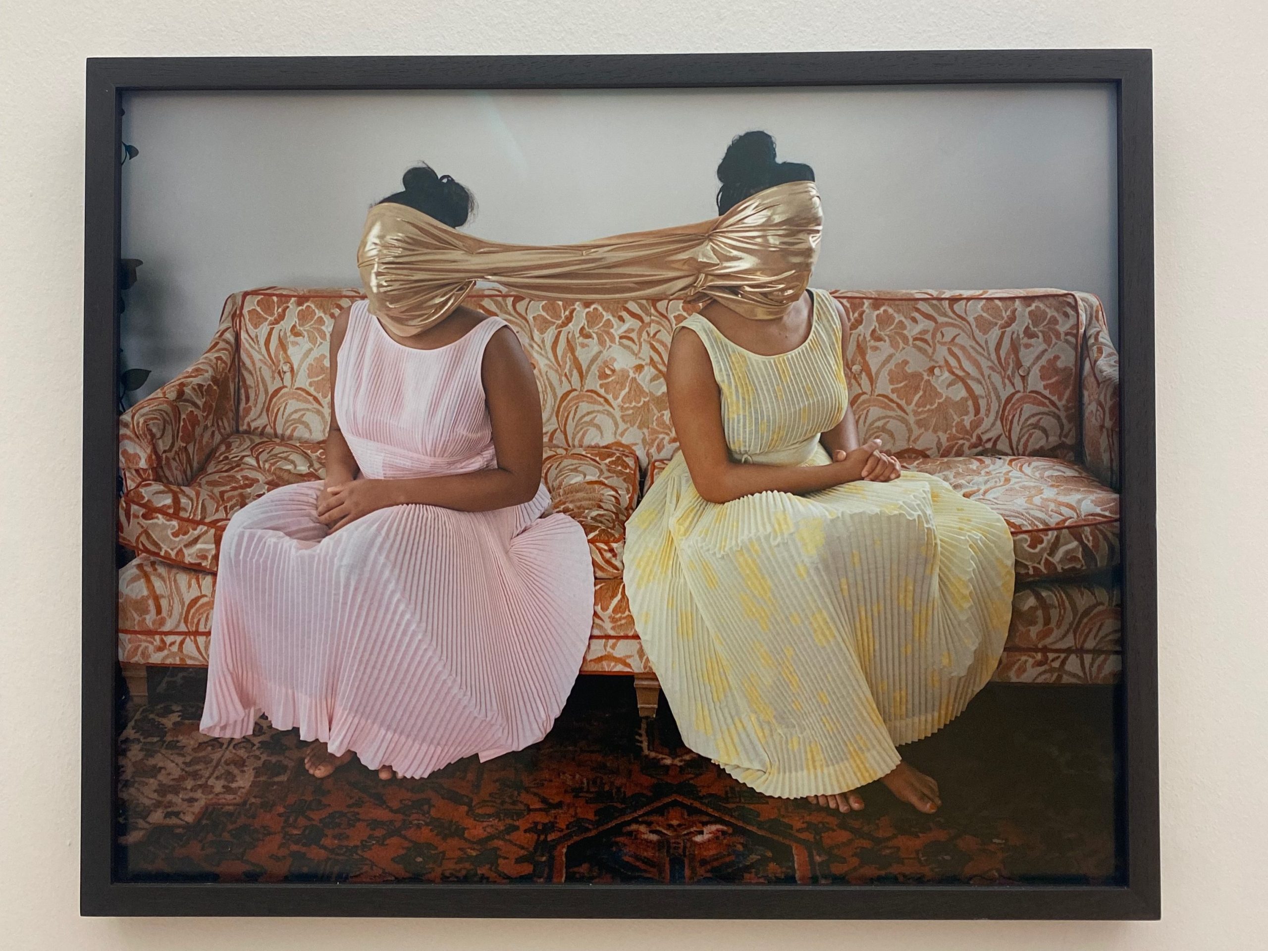 Image: Nydia Blas, "Gold The Girl Who Spun Gold." A framed photo of two Black women seated on a couch. Their posture and body language is identical: hands folded in their laps, feet crossed, hair up in a bun, at a 45 degree angle view away from the viewer, but in opposite directions, turned from each other. The one on the right wears a yellow translucent dress, while the one on the left wears a pink translucent dress. Connecting the two of them is a brassy gold cloth stretched between their heads, covering their faces. Photo by Jasmine Barnes.