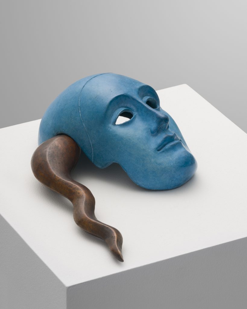 Image: Sif Itona Westerberg, Crux, 2022. Image credit: courtesy of the artist and Zinck Editions, Denmark. A blue mask is presented on a white plinth. The empty eye sockets look into the upper right corner of the image. A bronze, squiggly form emerges from the left side of the mask, ending in a point near the edge of the plinth. 