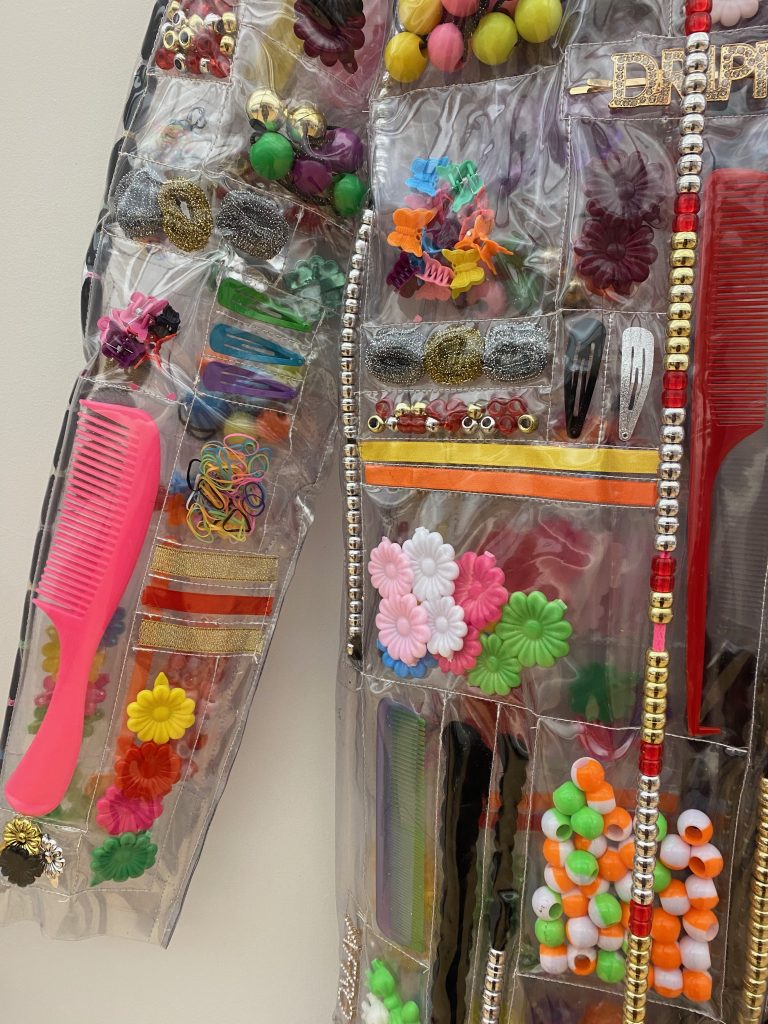 Image: A close up of Shaquita Reed's "For the Culture Product Jacket" (2021). The sleeve and torso of a transparent plastic jacket. Sewn into the jacket in discreet pockets are a variety of colorful objects such as beads, a pink comb, plastic flowers, rubber bands, hair barrettes,  and hair clips. Photo by Jasmine Barnes.