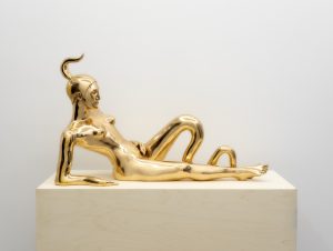 Image: Sif Itona Westerberg, "Ascendance," 2023. Image credit: courtesy of the artist and Gether Contemporary, Copenhagen. A golden figure reclines on a light wooden plinth. The figure's left leg is a smooth, undulating tendril while its right leg extends straight and pointed towards the edge of the plinth. The figure's right arm props it up and is also smooth, lacking definition. A single, smooth horn curves upwards from the figure's head.