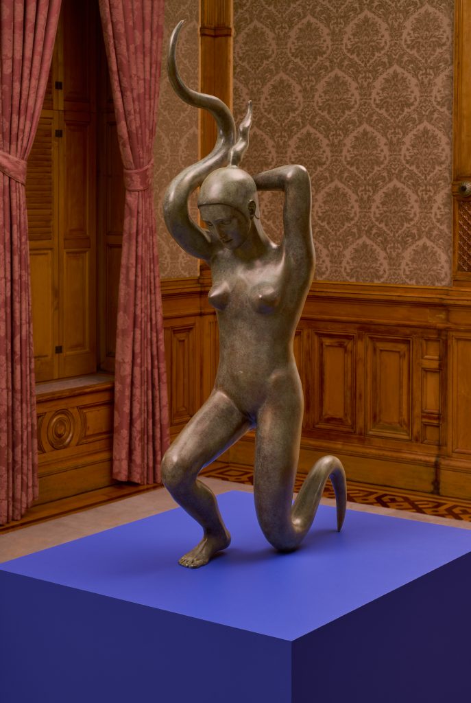 Image: Sif Itona Westerberg, Surrender, 2022/23/24. Image credit: Brian Griffin. A bronze figure is presented on a blue plinth. She is kneeling, her face downturned, and her left leg and right arm are smooth, wavy, and tendril-like. Her left arm is positioned behind her head with her hand pointing down the length of her spine. 