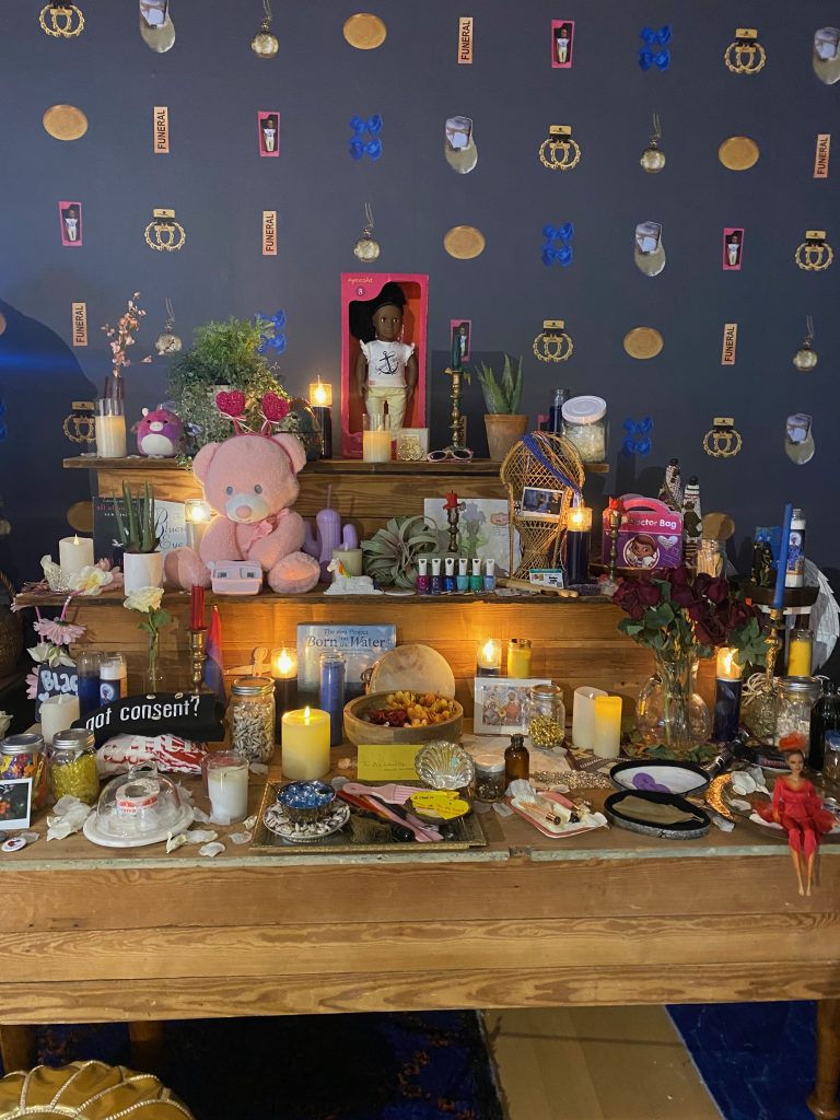 Image: "The Black Girlhood Altar, A Long Walk Home" 2023. Atop three wooden shelves, which get sequentially smaller, are a variety of objects, including hairpicks, incense, roses, a pink teddy bear, a Black barbie doll in a red dress, nail polish, a small wicker chair, and a lot of lit candles. At the top level, there is a Black doll in a pink box next to a snake plant. Photo by Jasmine Barnes.
